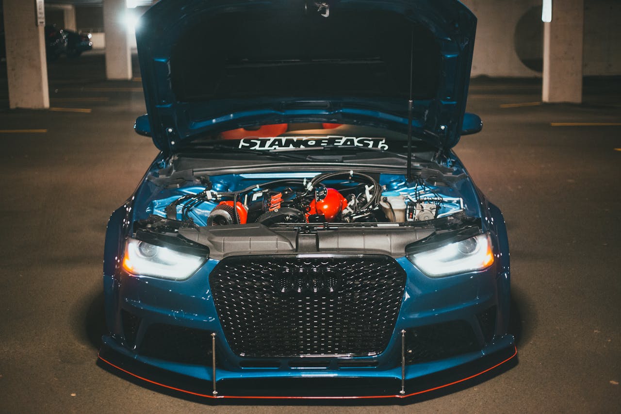 Home Blue sports car with open hood displaying engine in dimly lit urban setting.